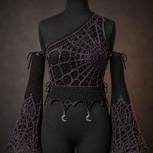 Könnte beinhalten: Ein schwarz-violettes Häkeloberteil mit asymmetrischem One-Shoulder-Ausschnitt und langen, ausgestellten Ärmeln mit Spinnennetzmuster. Das Kleidungsstück hat eine taillierte Passform mit spitzem Saum und zwei silberfarbenen Mondsichel-Anhängern.