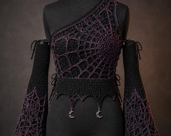 May include: A black and purple crochet top featuring an asymmetrical one-shoulder neckline and long, flared sleeves with a spiderweb pattern. The garment includes a fitted waist with a pointed hem and two silver-toned crescent moon charms.