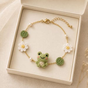 May include: A gold-tone chain bracelet featuring a central crocheted green frog, two white crocheted daisy charms, two green crocheted lily pad charms, and small white faux pearls. The bracelet includes a lobster claw clasp and an extender chain.