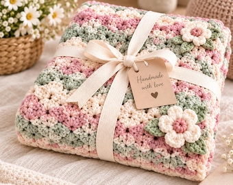 May include: A folded crochet blanket featuring a floral pattern in shades of pink, sage green, and cream. The blanket is tied with a cream-colored ribbon and a rectangular brown paper tag with the text Handmade with love and a heart icon.