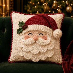 May include: A square crochet pillow featuring a Santa Claus face with a red hat, white beard, and mustache. The pillow is cream-colored with a dark red pom-pom border. A green holly sprig with red berries is attached to the side of the hat.