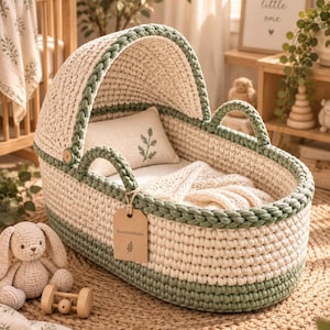 May include: A cream and sage green crocheted baby bassinet with a matching hood, two handles, and a small rectangular pillow featuring a green leaf embroidery. A brown paper tag with the text KnotStichStudio and a leaf icon hangs from one handle.