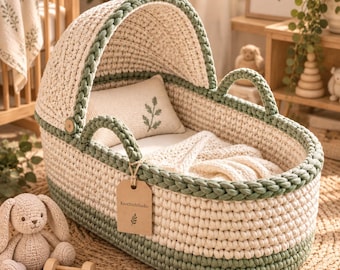 May include: A cream and sage green crocheted baby bassinet with a matching hood, two handles, and a small rectangular pillow featuring a green leaf embroidery. A brown paper tag with the text KnotStichStudio and a leaf icon hangs from one handle.