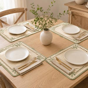 May include: Four rectangular placemats featuring a woven beige center and a decorative sage green crocheted border with floral motifs. Each placemat is set with a white ceramic plate, a folded beige napkin, and gold-toned flatware.