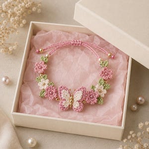 May include: A handmade crochet bracelet featuring a central butterfly motif in pink and cream, flanked by small pink and white flowers with green leaves. The bracelet has a pink cord with gold-toned beads and is presented in a square gift box.
