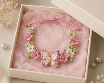 May include: A handmade crochet bracelet featuring a central butterfly motif in pink and cream, flanked by small pink and white flowers with green leaves. The bracelet has a pink cord with gold-toned beads and is presented in a square gift box.