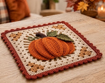 May include: A square crochet coaster featuring a central orange pumpkin with green leaves and a brown stem. The background is cream-colored with a decorative openwork pattern, accented by small brown leaves in the corners and a dark red scalloped border.
