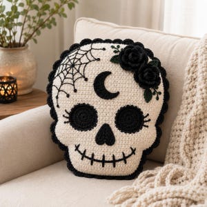 Könnte beinhalten: Ein cremefarbenes Häkelkissen in Totenkopfform mit schwarzen Garn-Details. Das Design zeigt einen schwarzen Halbmond auf der Stirn, ein Spinnennetz auf der linken Seite, zwei schwarze gehäkelte Rosen auf der rechten Seite sowie schwarz gestickte Augenhöhlen und einen Mund.
