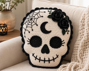 May include: A cream-colored crochet skull pillow with black yarn details. The design features a black crescent moon on the forehead, a spiderweb on the left side, two black crocheted roses on the right, and black stitched eye sockets and mouth.