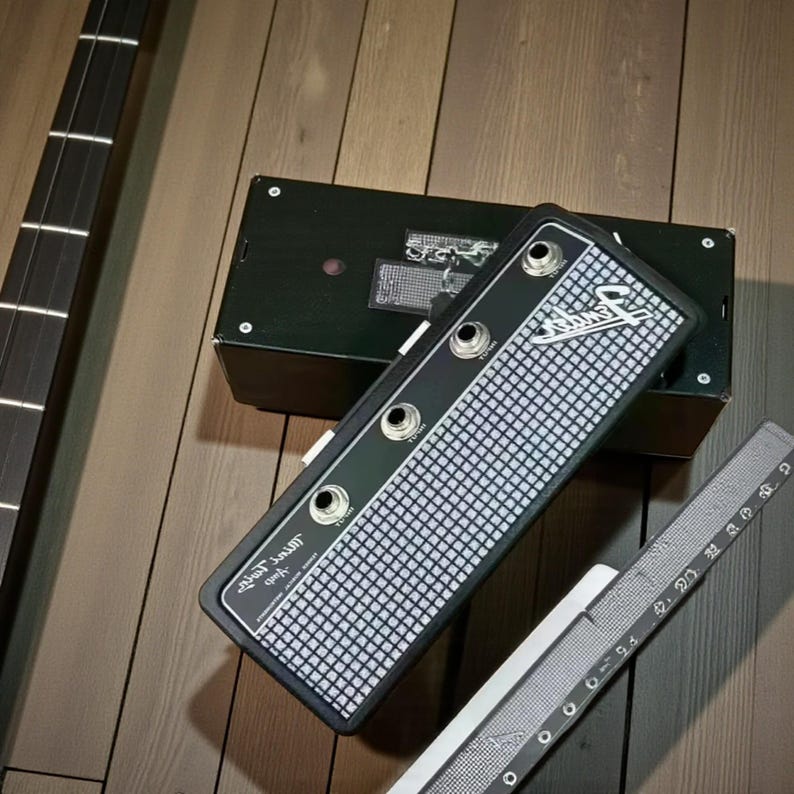 May include: A black Fender Mini Twin amplifier with a silver grid pattern front panel, featuring three input jacks and the Fender logo. A metal ruler is positioned alongside the device, showing a length of approximately 9.5 inches.