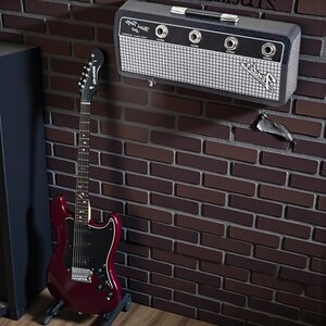 May include: A dark red electric guitar with a black pickguard stands on a floor stand next to a wall-mounted black and silver Fender Mini Twin amp featuring four input jacks and a white mesh grille with the Fender logo printed on the front.