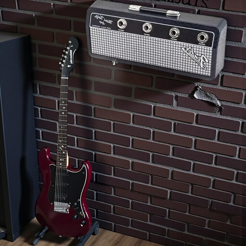 May include: A dark red electric guitar with a black pickguard stands on a floor stand next to a wall-mounted black and silver Fender Mini Twin amp featuring four input jacks and a white mesh grille with the Fender logo printed on the front.