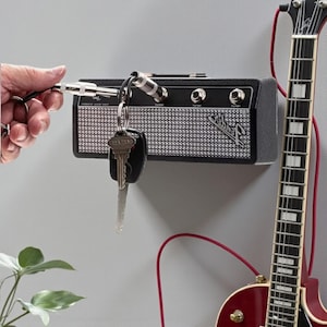 May include: A black wall-mounted key holder designed to resemble a miniature guitar amplifier with a silver mesh front and the Fender logo. It features four input jacks that serve as key hooks, with a metal plug keychain inserted into one jack.