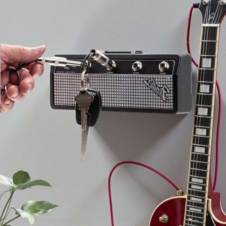 May include: A black wall-mounted key holder designed to resemble a miniature guitar amplifier with a silver mesh front and the Fender logo. It features four input jacks that serve as key hooks, with a metal plug keychain inserted into one jack.