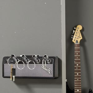 May include: A black wall-mounted key holder designed to resemble a guitar amplifier, featuring four input jacks that serve as key hooks. The front panel has a silver mesh grille, the Fender logo, and four metal key rings attached to the jacks.