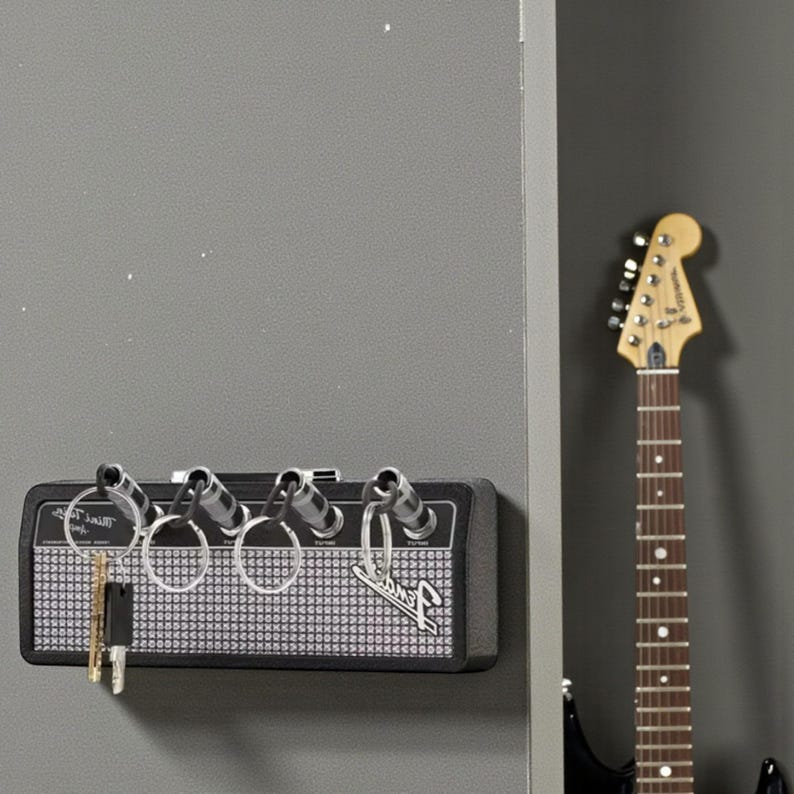 May include: A black wall-mounted key holder designed to resemble a guitar amplifier, featuring four input jacks that serve as key hooks. The front panel has a silver mesh grille, the Fender logo, and four metal key rings attached to the jacks.