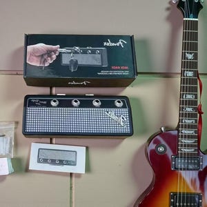 May include: A Fender Jack Rack wall-mounted key holder shaped like a guitar amplifier, featuring a black and silver mesh front with four input jacks. The product is shown in its black packaging box and includes a small instruction card nearby.