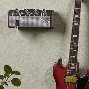 May include: A black wall-mounted key holder designed to resemble a guitar amplifier with a silver grille and the Fender logo. It features four input jacks that serve as key hooks, with four silver keychains inserted into the jacks.