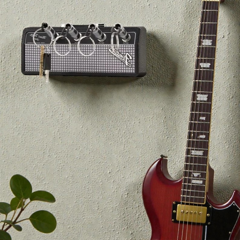 May include: A black wall-mounted key holder designed to resemble a guitar amplifier with a silver grille and the Fender logo. It features four input jacks that serve as key hooks, with four silver keychains inserted into the jacks.