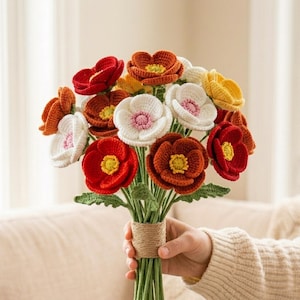 May include: A bouquet of crocheted flowers featuring red, orange, white, and yellow petals with textured centers. The stems are green and bound together with a thick, tan twine wrap, creating a structured arrangement of handcrafted textile blooms.