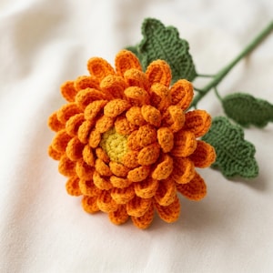 May include: A handcrafted crochet flower featuring vibrant orange petals arranged in multiple layers around a small, textured yellow centre. The flower is attached to a thin green stem with three distinct, serrated green leaves extending outwards.