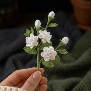 Può includere: Un ramo di fiori di gelsomino lavorato all'uncinetto a mano con tre fiori bianchi completamente sbocciati e due boccioli chiusi. Il ramo include diverse foglie verdi strutturate con dettagli a punto visibili, fissate su un sottile stelo di filo verde.