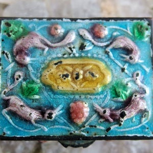 May include: A small, square, blue enamel box with a decorative design of pink and green stylized animals and flowers. The box has a gold-colored accent in the center.