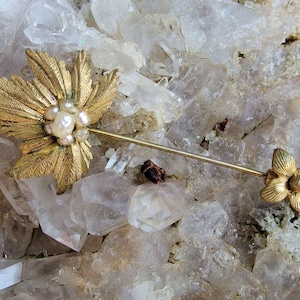 Puede incluir: Un broche dorado con un diseño de hoja y detalles de perlas. El broche está unido a un alfiler largo y delgado.