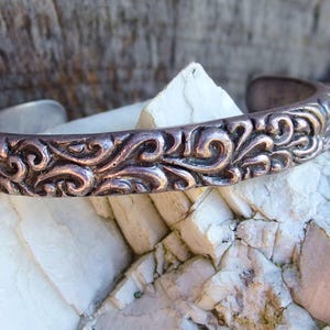 May include: A silver cuff bracelet featuring an intricate, swirling floral pattern. The bracelet has a slightly oxidised finish, giving it a vintage look. The open design allows for easy wear. The bracelet rests on a white and tan rock surface.