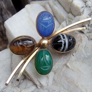 May include: A gold brooch with five scarab beetle shaped stones. The stones are blue, brown, green, and black and white.