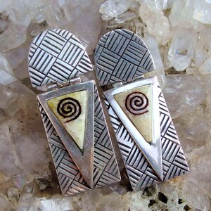 May include: A pair of silver earrings with a geometric design and a triangular inlay featuring a spiral pattern.