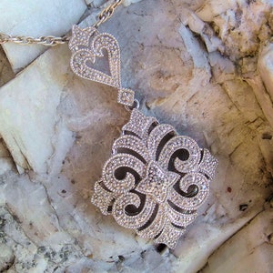 Può includere: Una collana con ciondolo in argento, con un designo dettagliato e ornato. Il ciondolo è di forma quadrata con un piccolo design a forma di cuore nella parte superiore. La collana è su una catena d'argento.