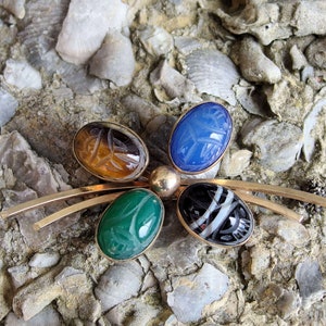 May include: A gold tone brooch with five carved gemstone cabochons. The stones are green, blue, brown, black and white. The brooch is shaped like a butterfly.