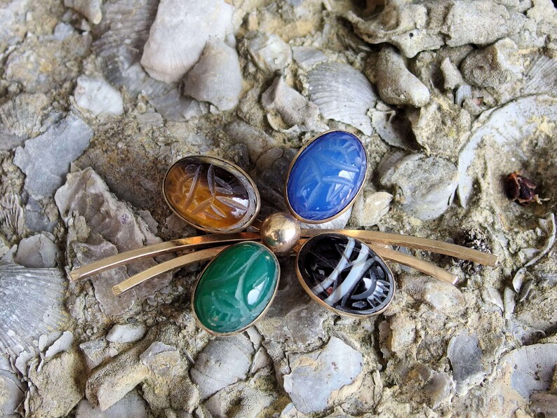 May include: A gold tone brooch with five carved gemstone cabochons. The stones are green, blue, brown, black and white. The brooch is shaped like a butterfly.