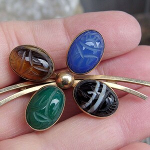 May include: A gold tone brooch with five carved gemstone cabochons. The stones are blue, green, brown, black and white. The brooch is in the shape of a stylized flower.