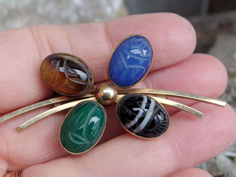 May include: A gold tone brooch with five carved gemstone cabochons. The stones are blue, green, brown, black and white. The brooch is in the shape of a stylized flower.