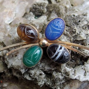 May include: A gold tone brooch with five oval shaped stones. The stones are carved with an Egyptian scarab beetle design. The stones are brown, blue, green, black and white.