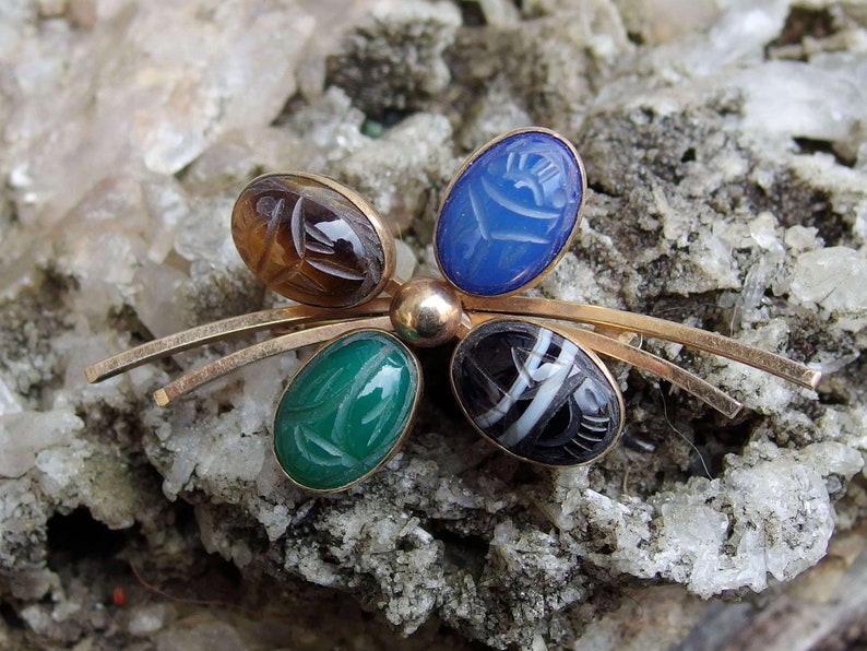 May include: A gold tone brooch with five oval shaped stones. The stones are carved with an Egyptian scarab beetle design. The stones are brown, blue, green, black and white.