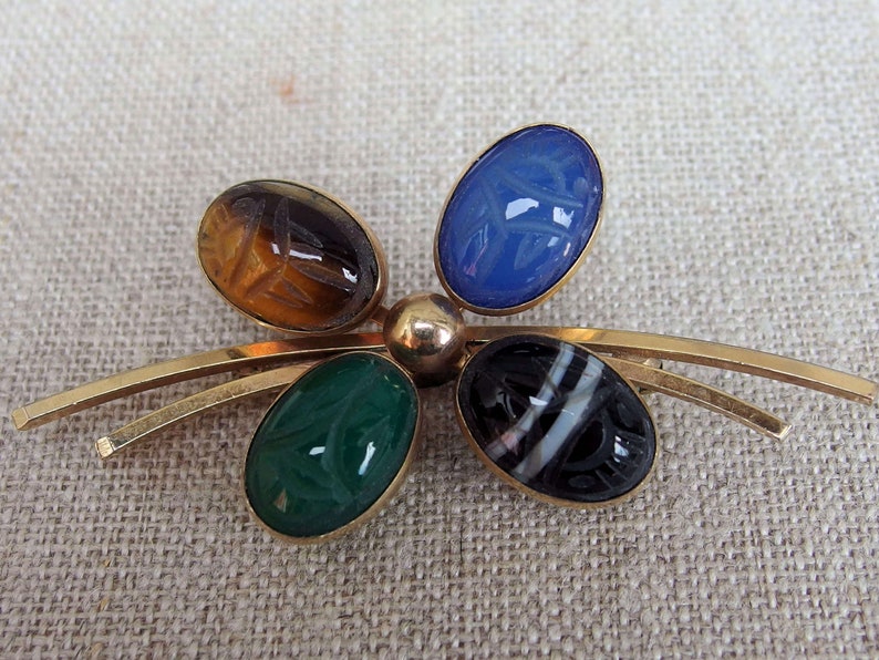 May include: A gold tone brooch with five oval shaped stones. The stones are carved with an Egyptian scarab beetle design. The stones are blue, green, brown, black and white.
