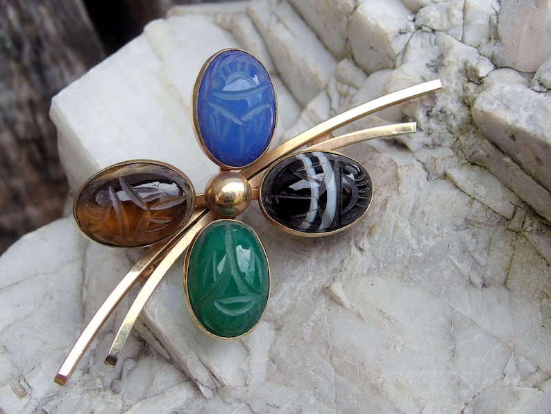 May include: A gold brooch with five scarab beetle shaped stones. The stones are blue, brown, green, and black and white.
