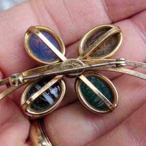 May include: Gold tone butterfly brooch with four oval shaped stones set in gold tone frames. The stones are green, blue, brown, and black and white striped. The brooch has a safety clasp and the words "BALTRON" are engraved on the back.