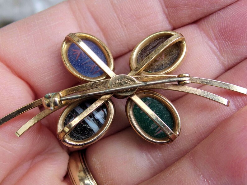 May include: Gold tone butterfly brooch with four oval shaped stones set in gold tone frames. The stones are green, blue, brown, and black and white striped. The brooch has a safety clasp and the words "BALTRON" are engraved on the back.