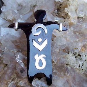 May include: A black pendant shaped like a person with outstretched arms, with silver-coloured detailing. The pendant has a glossy finish and is set against a background of light-coloured crystals. The silver accents include a spiral, a chevron, a dot, and a curved shape.