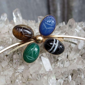 May include: A gold tone brooch with four carved gemstone scarab beetles. The beetles are green, blue, brown, and black. The brooch is set on a bed of white crystals.