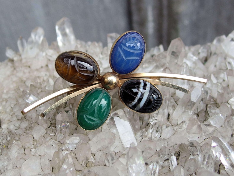 May include: A gold tone brooch with four carved gemstone scarab beetles. The beetles are green, blue, brown, and black. The brooch is set on a bed of white crystals.