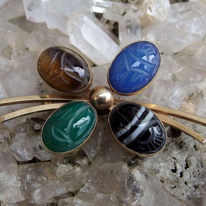 May include: A gold tone brooch with five oval shaped stones. The stones are carved with scarab beetle designs and are set in a gold tone metal. The stones are green, blue, brown, black and white.
