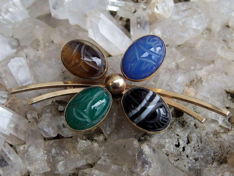 May include: A gold tone brooch with five oval shaped stones. The stones are carved with scarab beetle designs and are set in a gold tone metal. The stones are green, blue, brown, black and white.