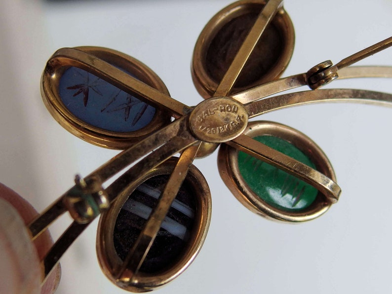May include: Gold tone brooch with four oval shaped cabochons. The cabochons are blue, green, brown, and white. The brooch has a hinged clasp and is marked "Val-Ron Jewelry".