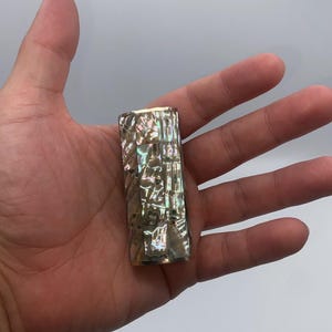 May include: A rectangular, iridescent abalone shell lighter. The lighter is held in an open hand against a white background. The shell has a textured surface with shades of green, blue, and gold.
