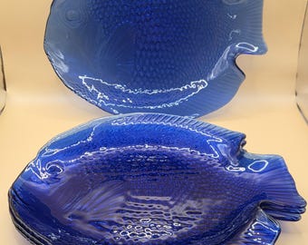 Op de afbeelding: Een set kobaltblauwe glazen borden in de vorm van een vis. De borden zijn voorzien van een gestructureerd schubbenpatroon op het lichaam, gedefinieerde vinnen en een duidelijk oogdetail. Het glas is doorschijnend en diepblauw, met één bord rechtop achter een stapel van drie identieke borden.